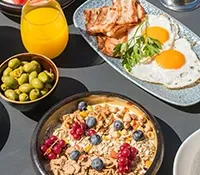 Breakfast spread with cereal bowl, eggs, bacon, olives, and orange juice.