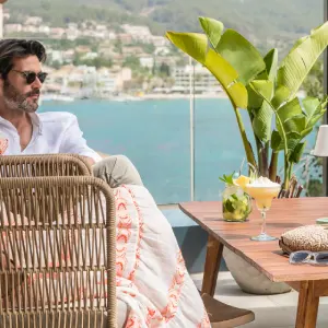 Man relaxing on patio with drink, sea view and plants in background.