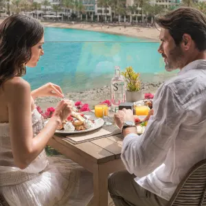 Couple dining with a seaside view, enjoying a meal with orange juice and a bottle of water.