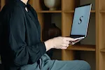 Person in black shirt holding a laptop with a letter "S" on the screen.