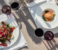 Two plates of pasta and salad with red wine on a table in sunlight.
