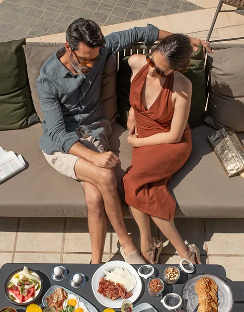 Couple sitting on a sofa outdoors with a table of assorted snacks and drinks in front of them.