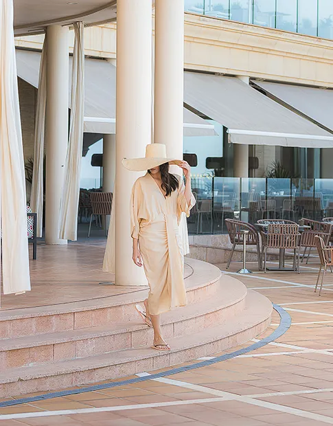 Woman in beige outfit and hat walks down steps in an outdoor patio area.
