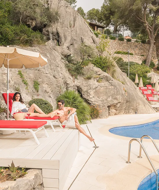 Couple relaxing on red lounge chairs by a pool with rocky backdrop.