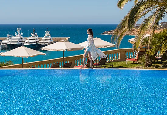 Woman by a poolside overlooking yachts and the ocean, with palm tree and umbrellas nearby.