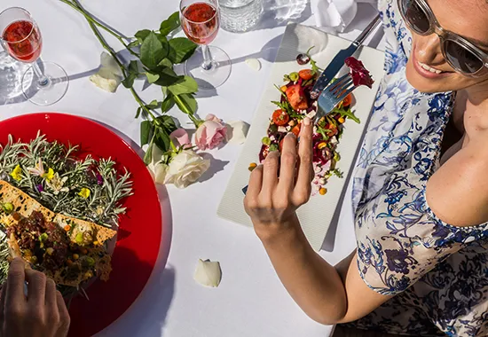Woman dining outdoors with elegant food and drinks, surrounded by roses on a white tablecloth.