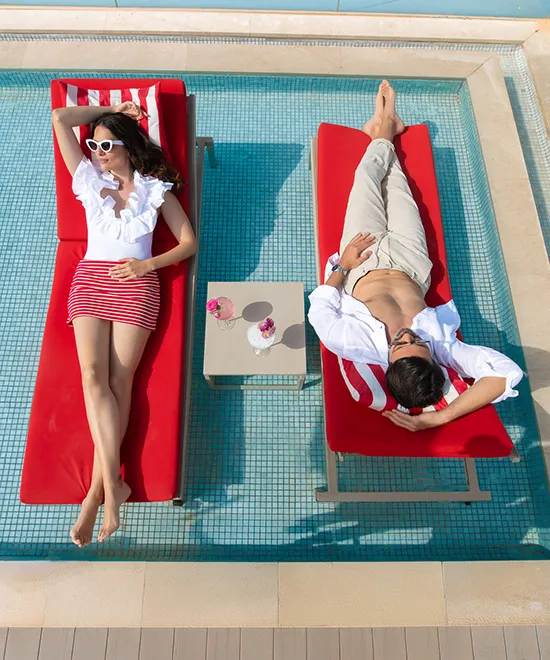Two people on red loungers beside a small table in a shallow pool, enjoying a sunny day.