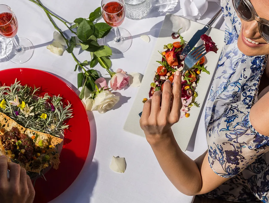 Woman enjoying a colorful salad at an outdoor table with roses and drinks.