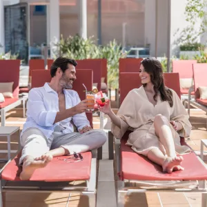 A couple toasting with drinks on red loungers at a poolside.