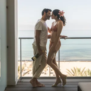 Couple on a balcony by the beach, with one holding a wine bottle, sharing a tender moment.