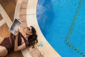 Woman by a pool reading a magazine, wearing sunglasses and a swimsuit with a drink beside her.