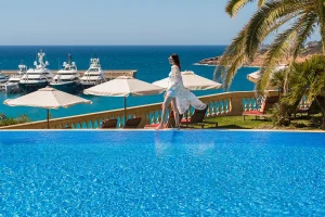 Woman by a pool with umbrellas, overlooking yachts and ocean under a clear blue sky.