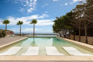 Pool with lounge chairs, palm trees, and a marina view under a blue sky.