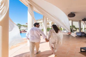 Couple holding hands and walking by a pool, with flowing curtains in a sunny setting.