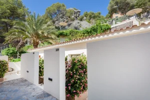 White building with a tiled roof, surrounded by lush greenery and flowers, under a clear blue sky.