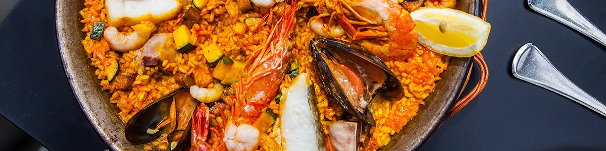 A pan of seafood paella with shrimp, mussels, and lemon wedges on a dark table.
