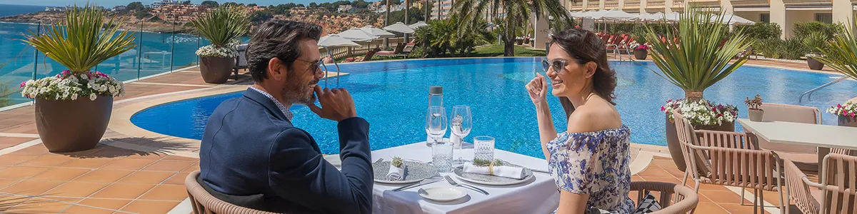 Couple dining by a pool with ocean view and sunny surroundings.