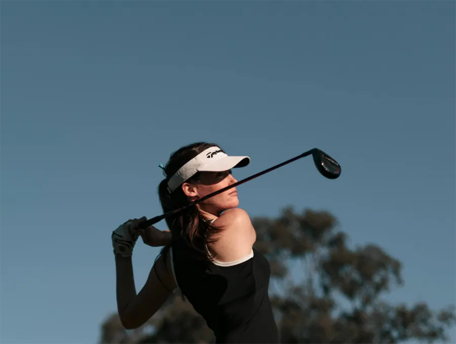 Golfer in a black dress and visor swinging a club against a blue sky background.