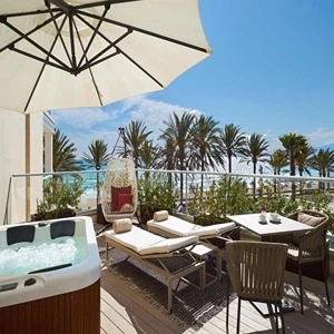 Sunny balcony with lounge chairs, hot tub, dining table, and palm trees in view.