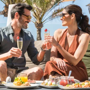 Couple enjoying drinks and food outdoors, with palm trees in the background.