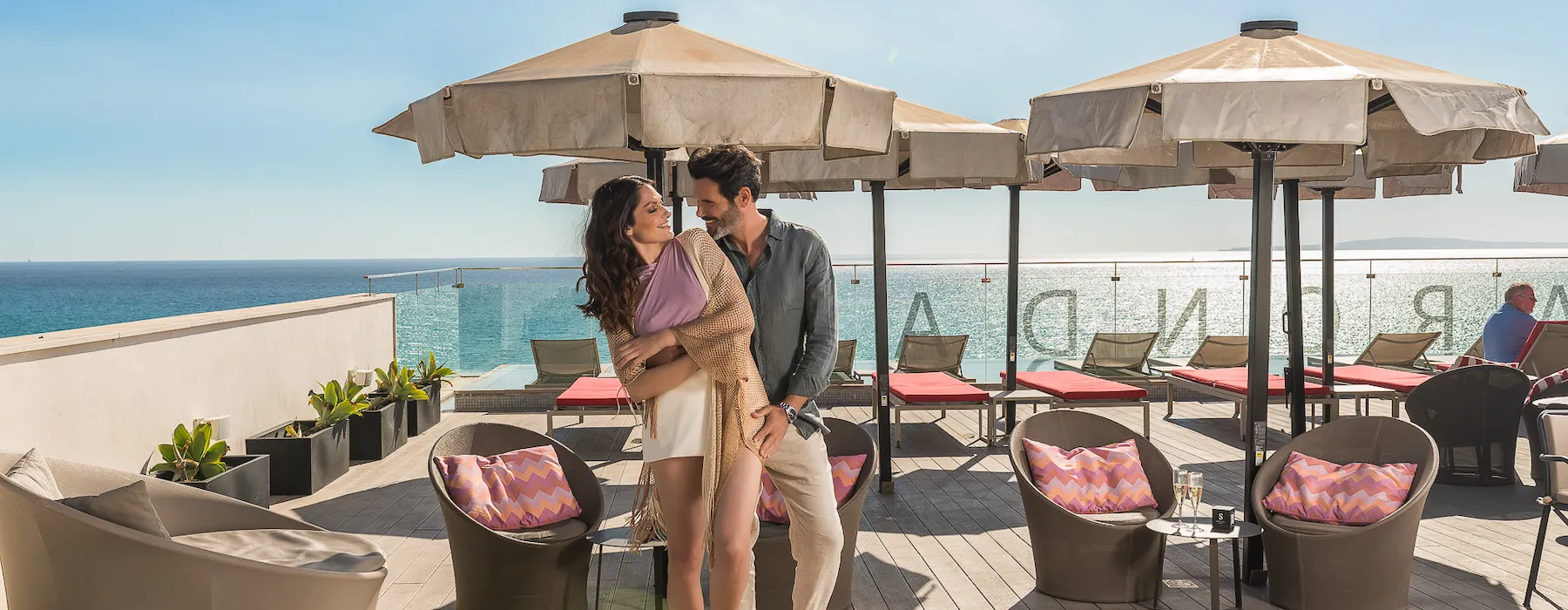 Couple smiling on a seaside terrace under umbrellas with seating and a clear blue sky.