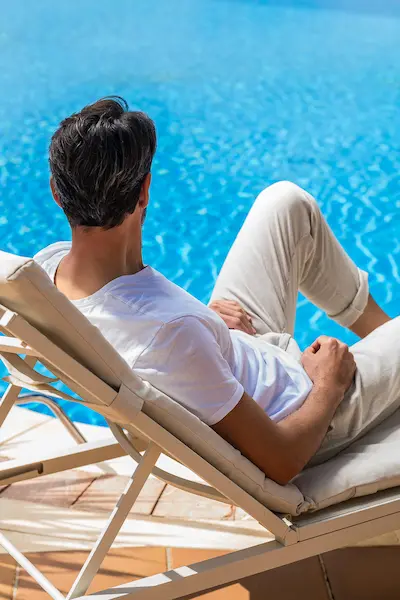 Man lounging in a deck chair by a bright blue pool in casual white attire.