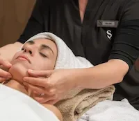 Person receiving a facial massage at a spa, wrapped in a towel, another person’s hands massaging their face.