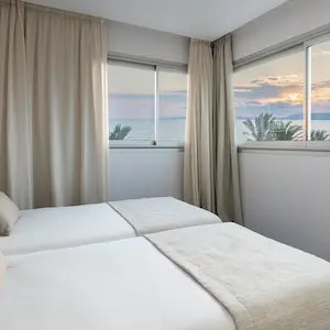 Bedroom with two beds, windows with curtains, and a sunset view over water and palm trees.