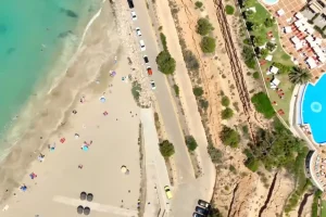 Aerial view of a beach with people, umbrellas, a road with parked cars, and a pool area on the right.