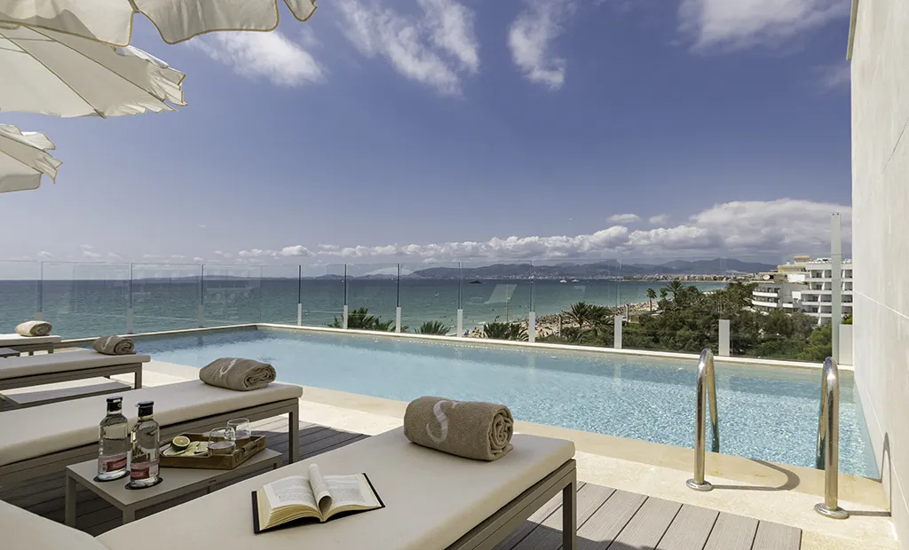 Sunlit poolside loungers with ocean view, towels, and a book under umbrellas.