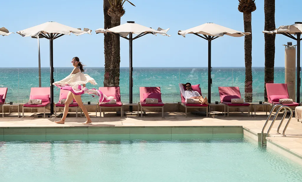 Poolside scene with pink loungers, umbrellas, and ocean view. Woman walking, another relaxing.