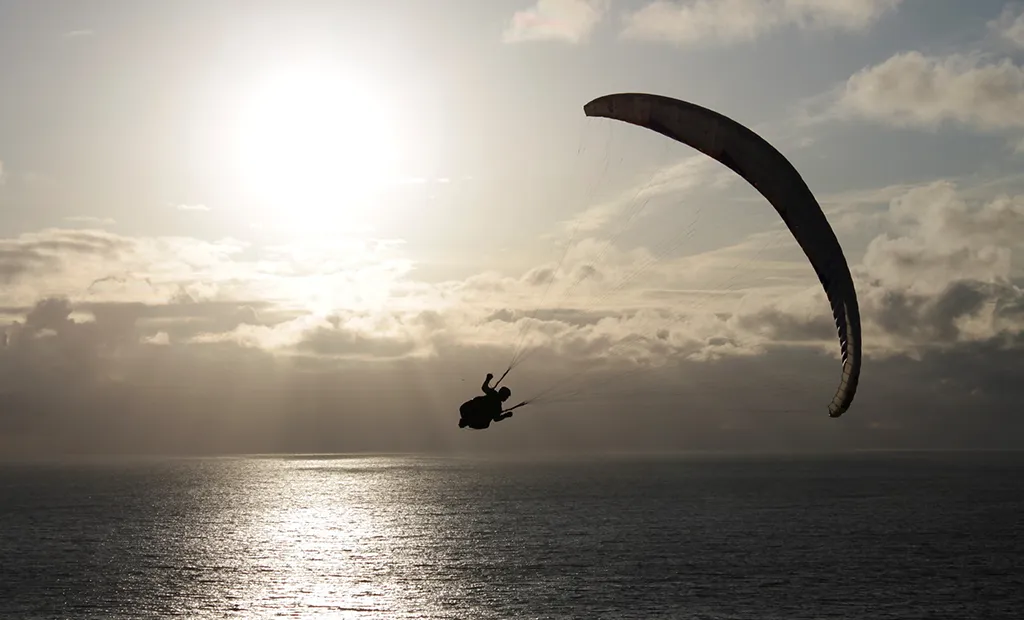 Paraglider soaring over the ocean with a dramatic cloudy sky and sun in the background.