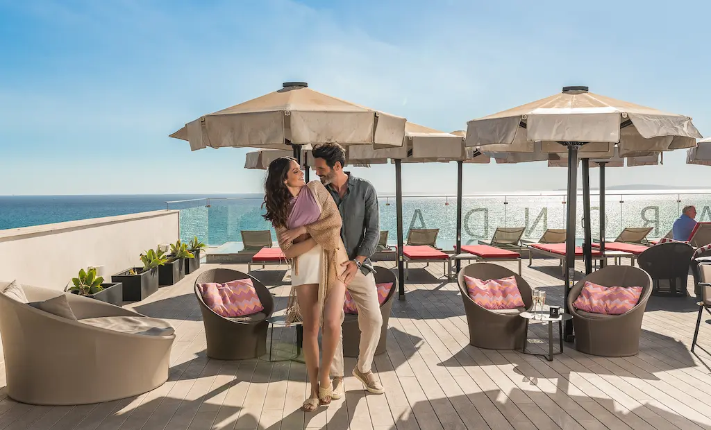 Couple smiling on a sunny deck with umbrellas, ocean view in the background.