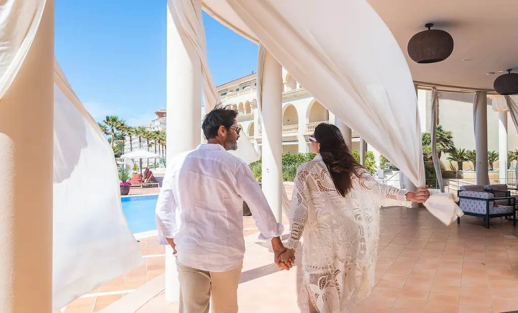 A couple in white, holding hands, walking near a poolside, with flowing curtains and sunny skies.