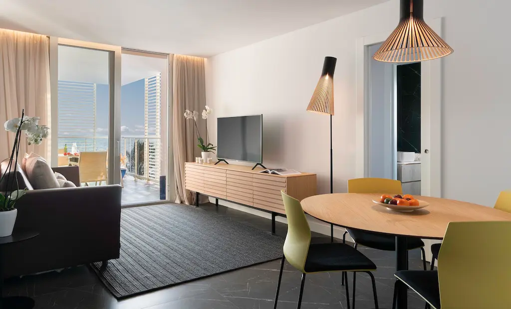 Modern living room with TV, round table, yellow chairs, balcony view, and large windows.
