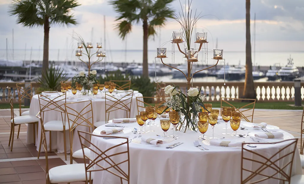 Elegant outdoor dining setup with white tablecloths, amber glasses, and a marina view.