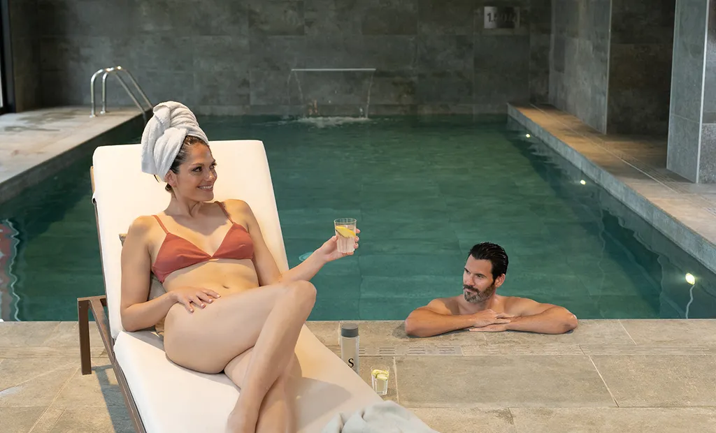 Man in pool beside woman relaxing on lounger with a drink, towel on her head.