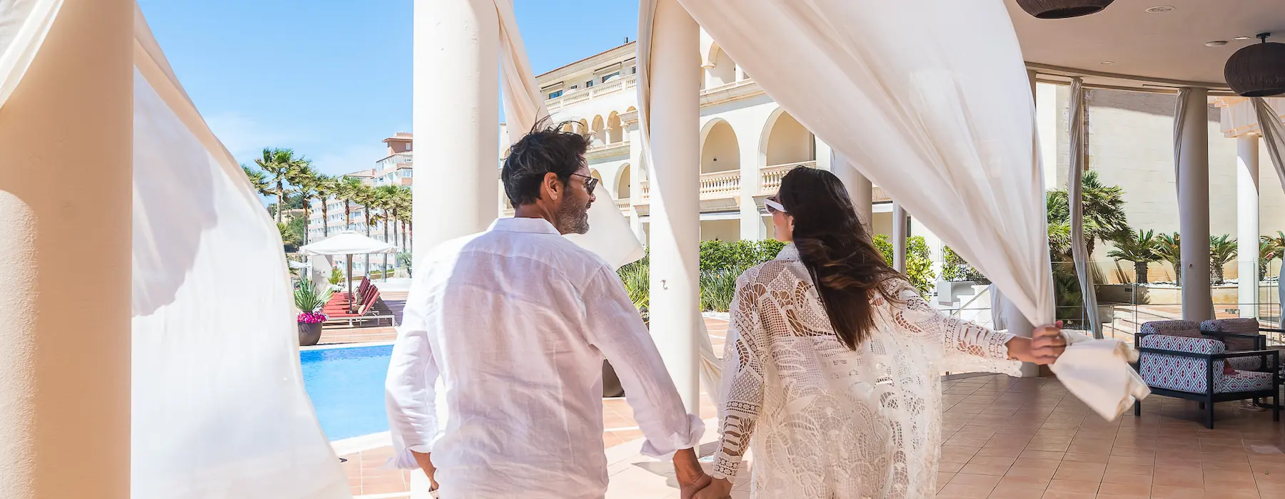 A couple holding hands near a pool with white drapes flowing in the breeze.