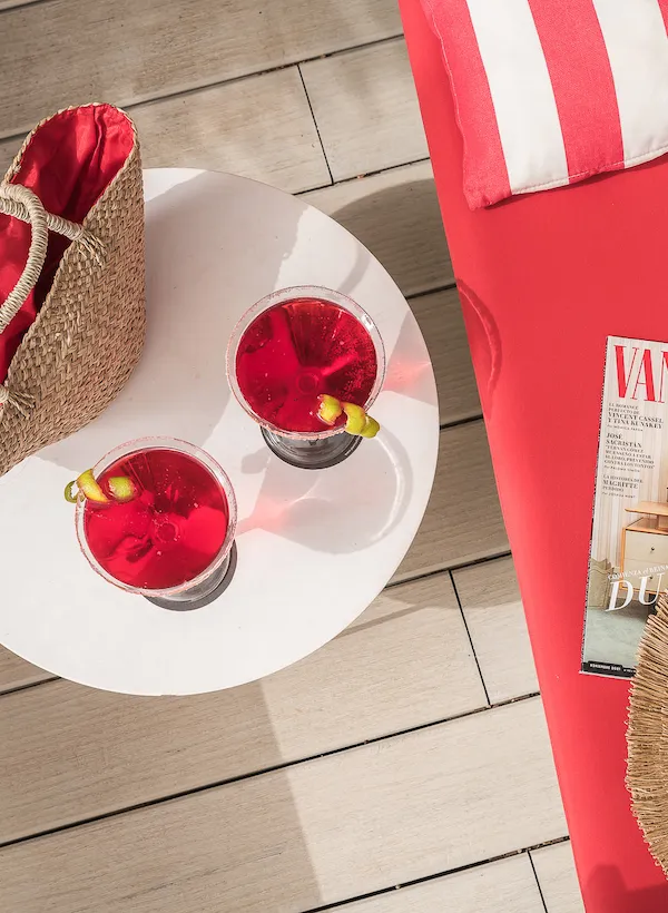 Two red drinks on a table next to a red lounge chair and a straw bag.