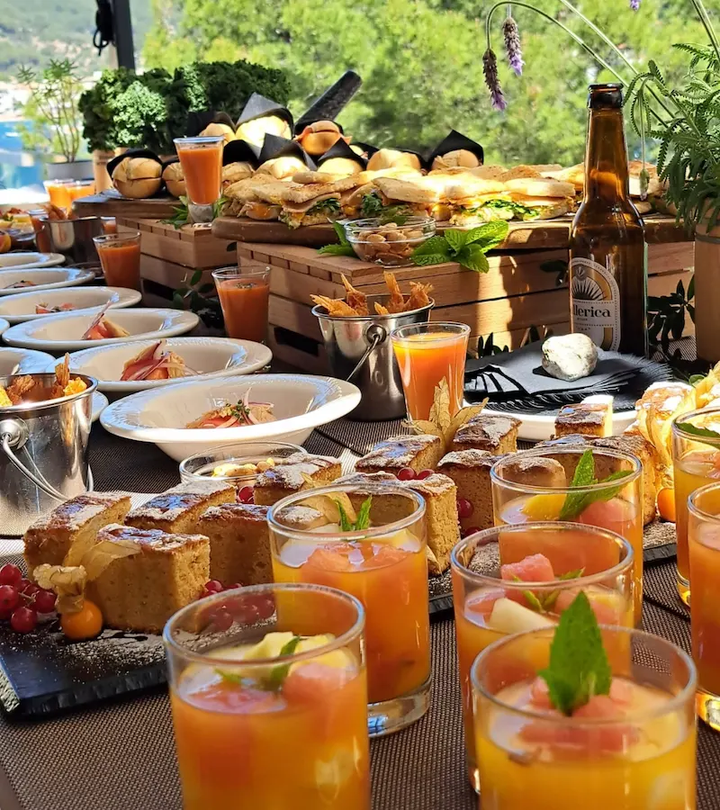Buffet table with various dishes, drinks, sandwiches, and desserts arranged outdoors.