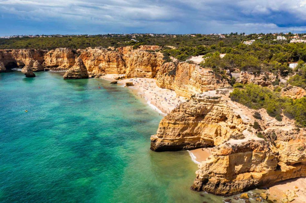 Aerial view of a beach surrounded by rocky cliffs and turquoise water, under a partly cloudy sky.