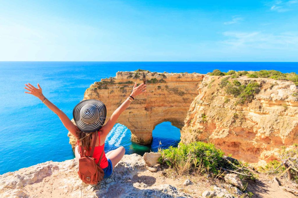 Person sitting on a cliff overlooking the ocean, arms raised, wearing a hat and backpack, vibrant sky and sea in the background.
