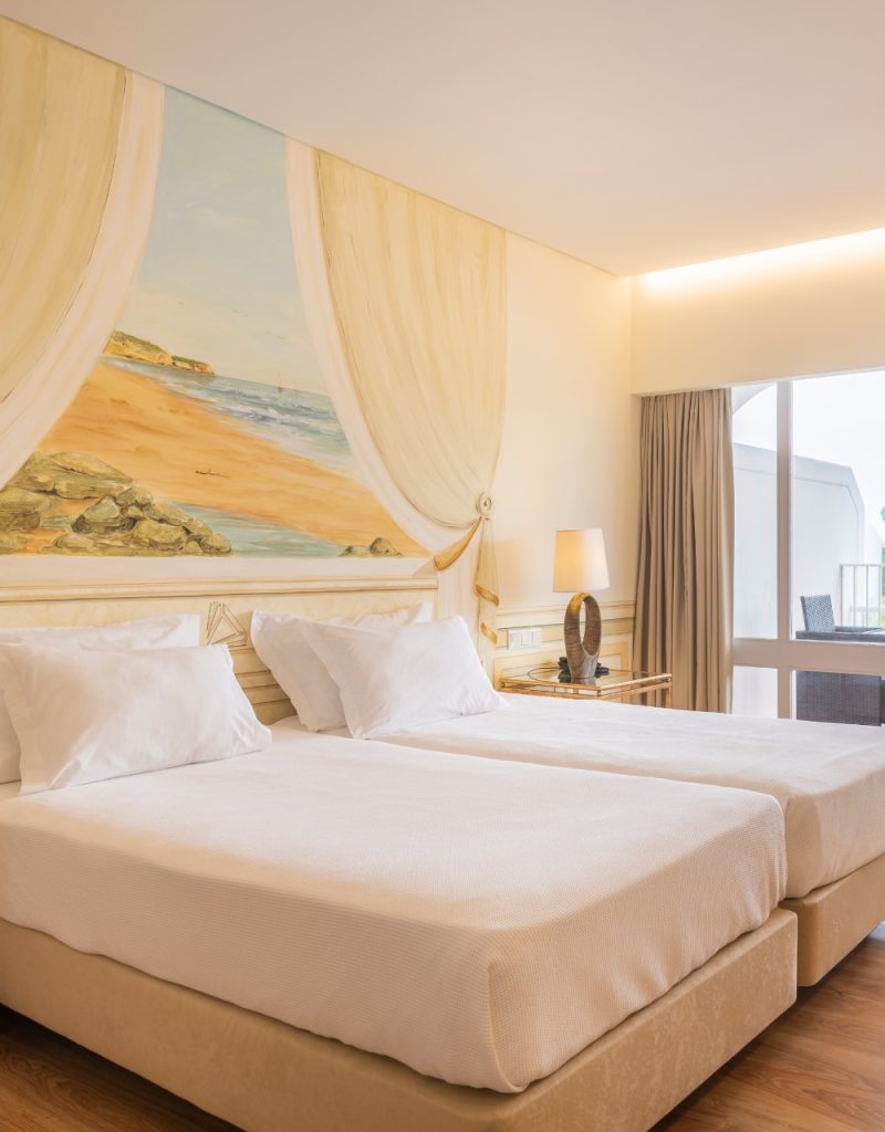 Two neatly made beds in a hotel room, with a beach mural on the wall and a lamp on a side table near a window.