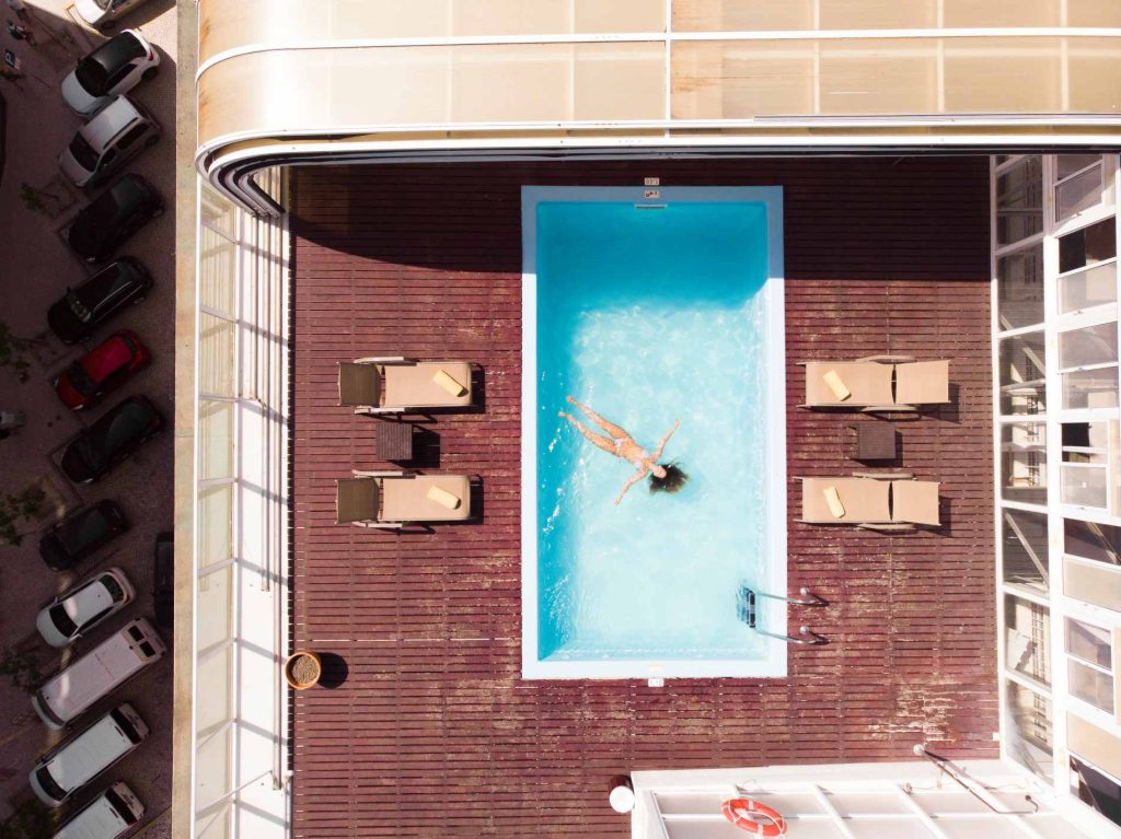 Aerial view of a person floating in a rooftop pool with lounge chairs and parked cars nearby.