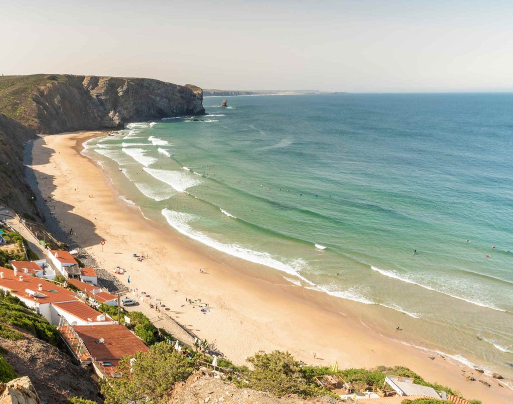 Aerial view of a beach with waves, cliffs, and people on the sand, with houses in the lower left corner.