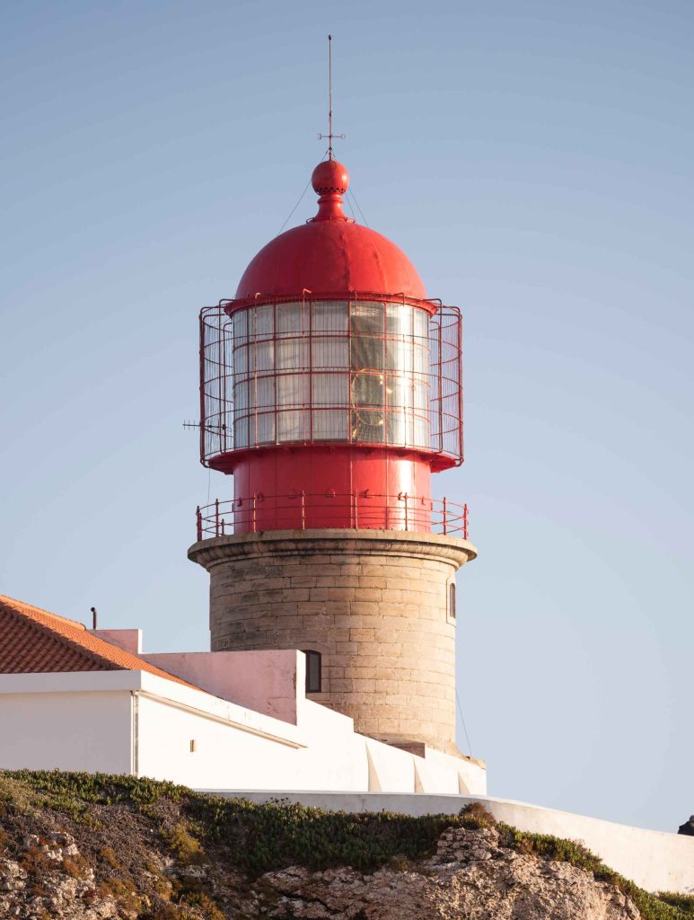 A red-domed lighthouse atop a stone structure with white buildings, set against a clear blue sky.