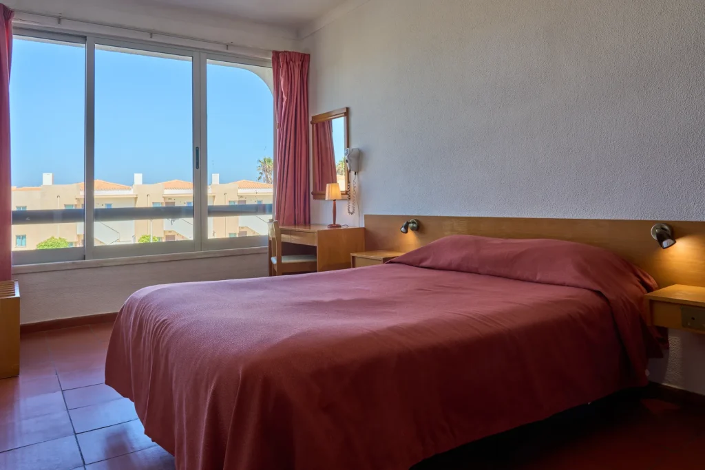Hotel room with a bed, red bedding, window view of rooftops, two lamps, a desk, and chair. Bright and tidy interior.