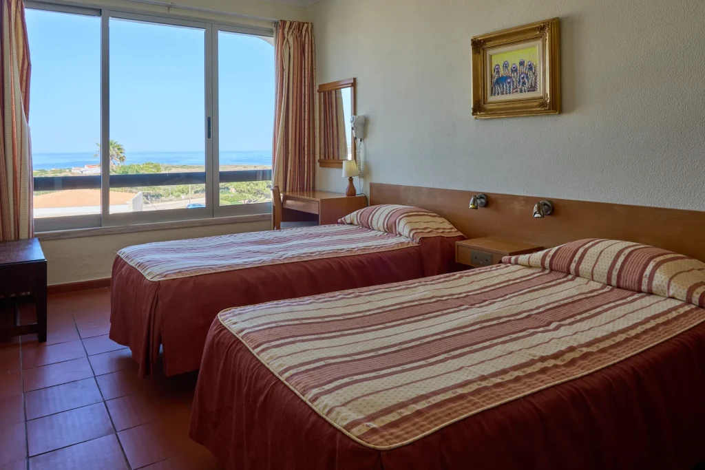 Hotel room with two striped beds, a painting on the wall, and a large window showing a sea view.