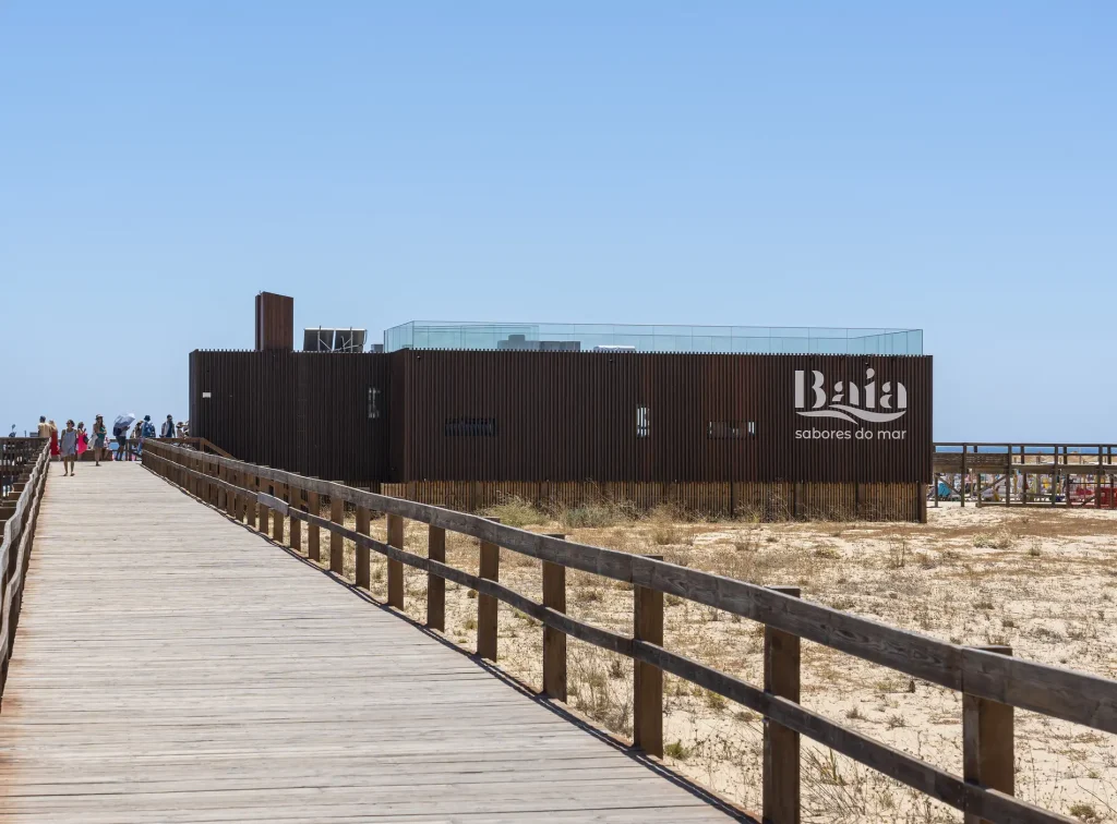 Wooden walkway leading to a modern beachside building with a clear blue sky.