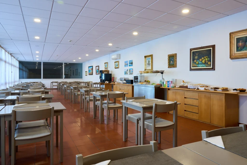 Empty cafeteria with wooden tables and chairs, framed art on walls, and food on side counter.